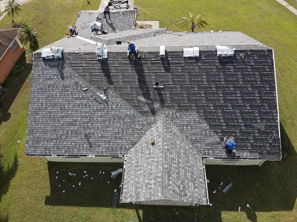 An aerial view captures workers diligently installing shingles on a house roof, surrounded by equipment and materials—a stark reminder of the intricate process and potential roof replacement costs involved.