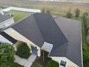 Aerial view of a single-story house with a dark shingle roof, often admired by roofing contractors, and a yellow exterior. It's surrounded by greenery and a white fence. The background reveals a misty landscape with trees and a road.