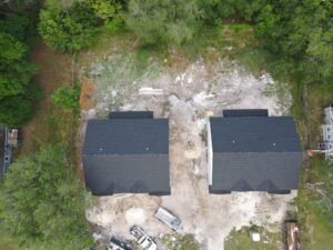 Aerial view of two unfinished houses with black roofs, surrounded by trees and construction materials, highlights the precision work typical of leading roofing companies in the area.