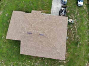 Aerial view of a house with a brown, shingled roof, expertly installed by roofing contractors. Two small skylights are visible. A driveway extends from the top right corner, where a parked vehicle is visible near a trailer with items. The surrounding yard is grassy and green.