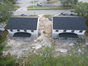 Aerial view of two two-story buildings under construction, with black roofs expertly laid by roofing contractors and unfinished exteriors. The site is cluttered with construction materials and debris. A road runs in front of the buildings, and greenery surrounds the area.