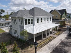 A two-story, light gray house with a metal roof installed by roofing contractors and a large front porch. It is surrounded by a small garden on a clear, sunny day. A streetlamp stands in the foreground, with neighboring houses visible in the background.