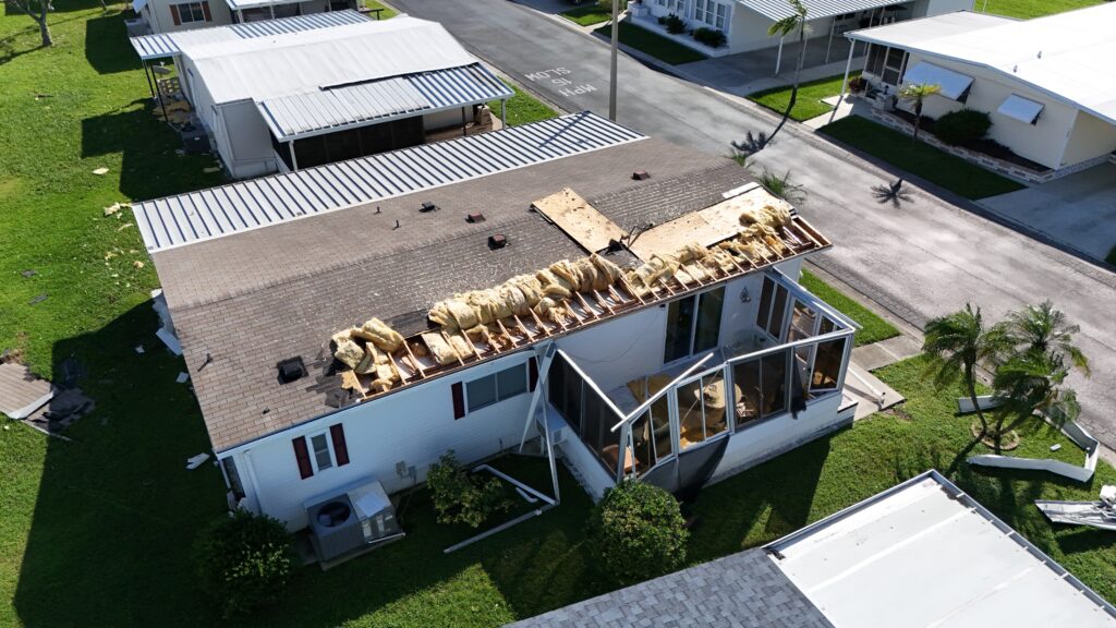 Aerial view of a house with roof damage, debris scattered, and surrounding homes intact highlights the urgent need for mobile home roof repair.