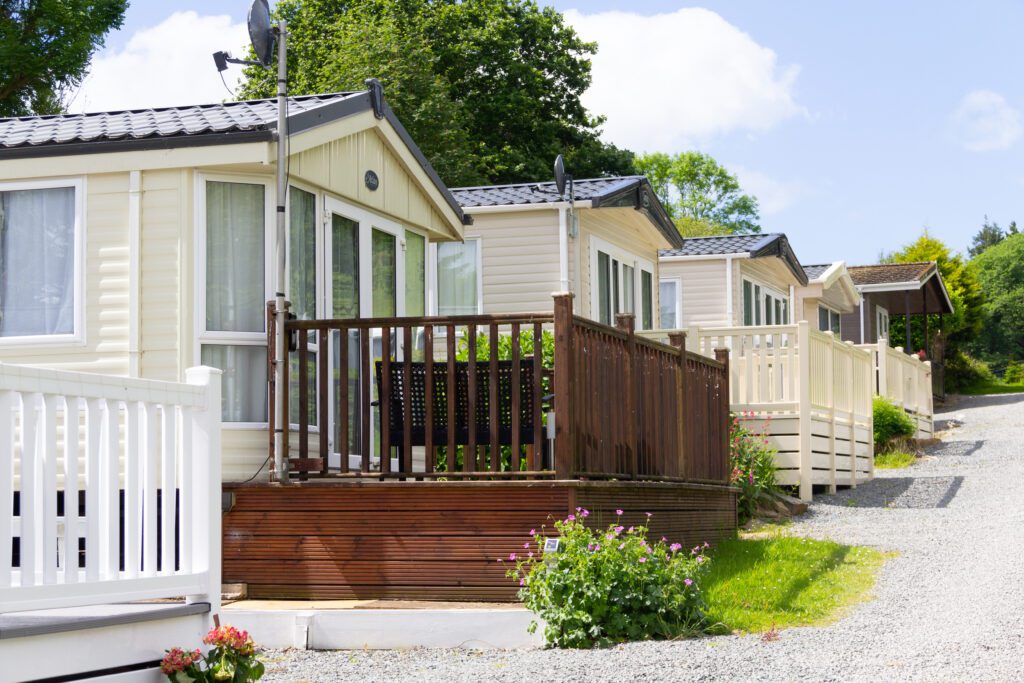 A row of mobile homes with wooden decks basks in the sun, surrounded by lush greenery and a gravel path, showcasing recent mobile home roof repairs that blend seamlessly into the serene setting.