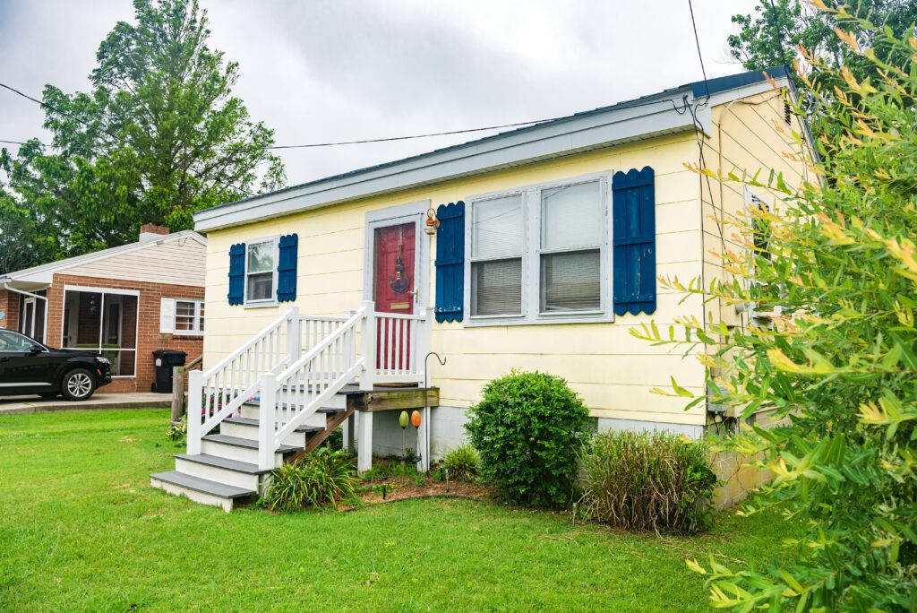 A small yellow house with blue shutters, front porch, and steps sits under a sturdy mobile home roof repair job. Surrounded by lush greenery on a cloudy day, this charming abode invites you in with its quaint appeal.