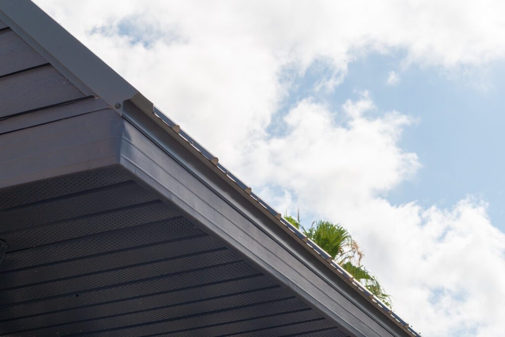 Close-up of a black roof corner featuring a roof drip edge, set against a cloudy blue sky and lush palm foliage in the background.