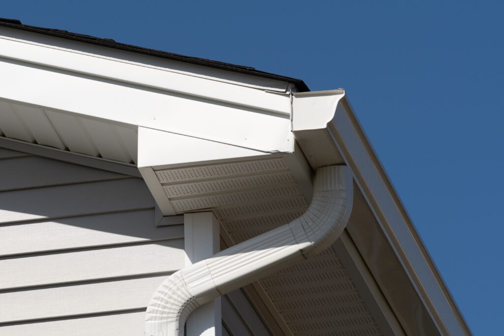 Close-up of a house roof corner showcasing the white gutters and downspout, with a neatly installed roof drip edge, all set against a clear blue sky.