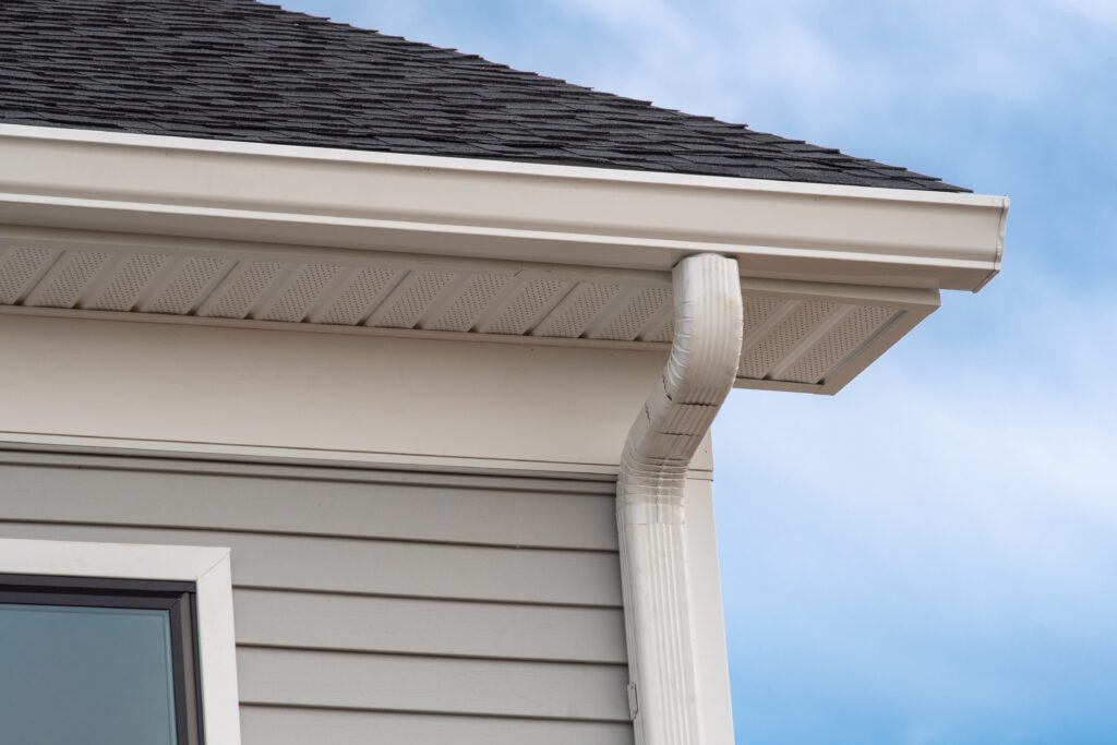 Close-up of a house roof corner featuring a gutter, downspout, and roof drip edge against a clear blue sky.