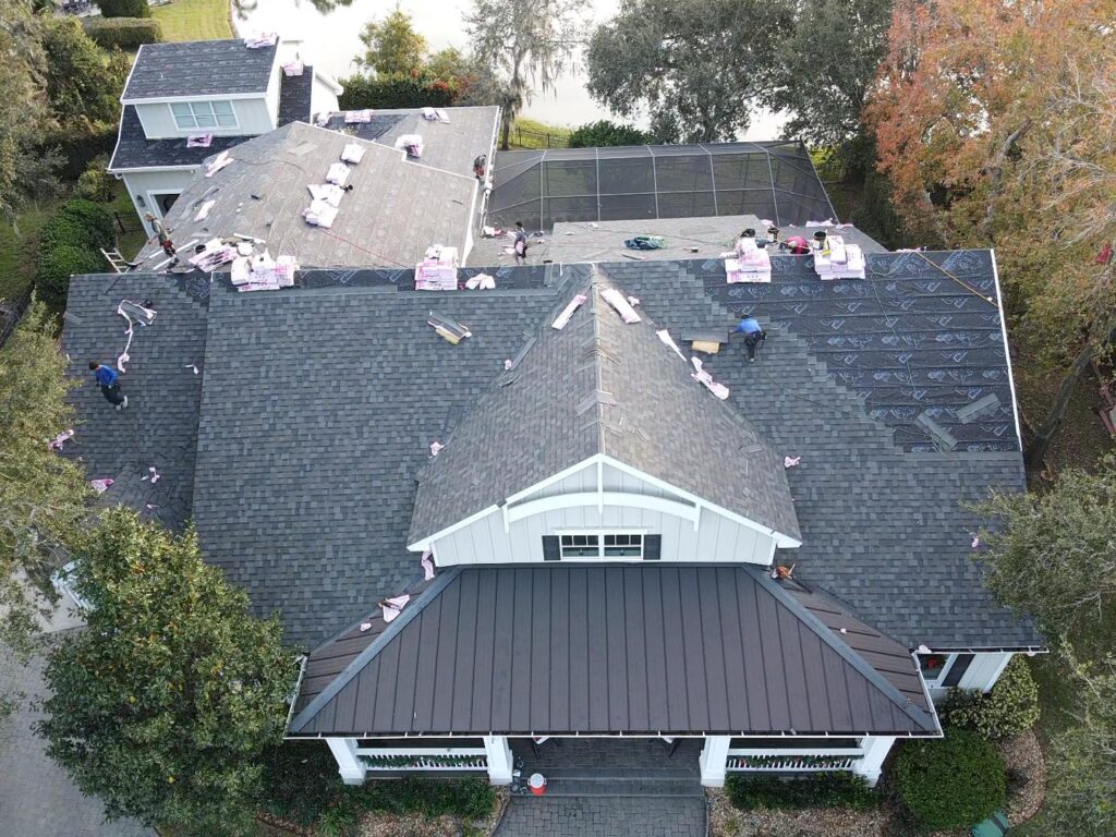 Aerial view of roofing contractors expertly installing shingles as part of a roof replacement on a large house surrounded by lush trees.