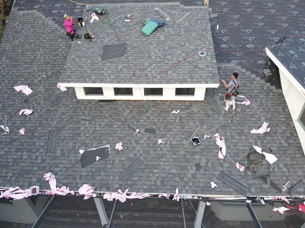 Roofers conducting a roof installation on a gray shingled roof, surrounded by tools and materials, viewed from above.