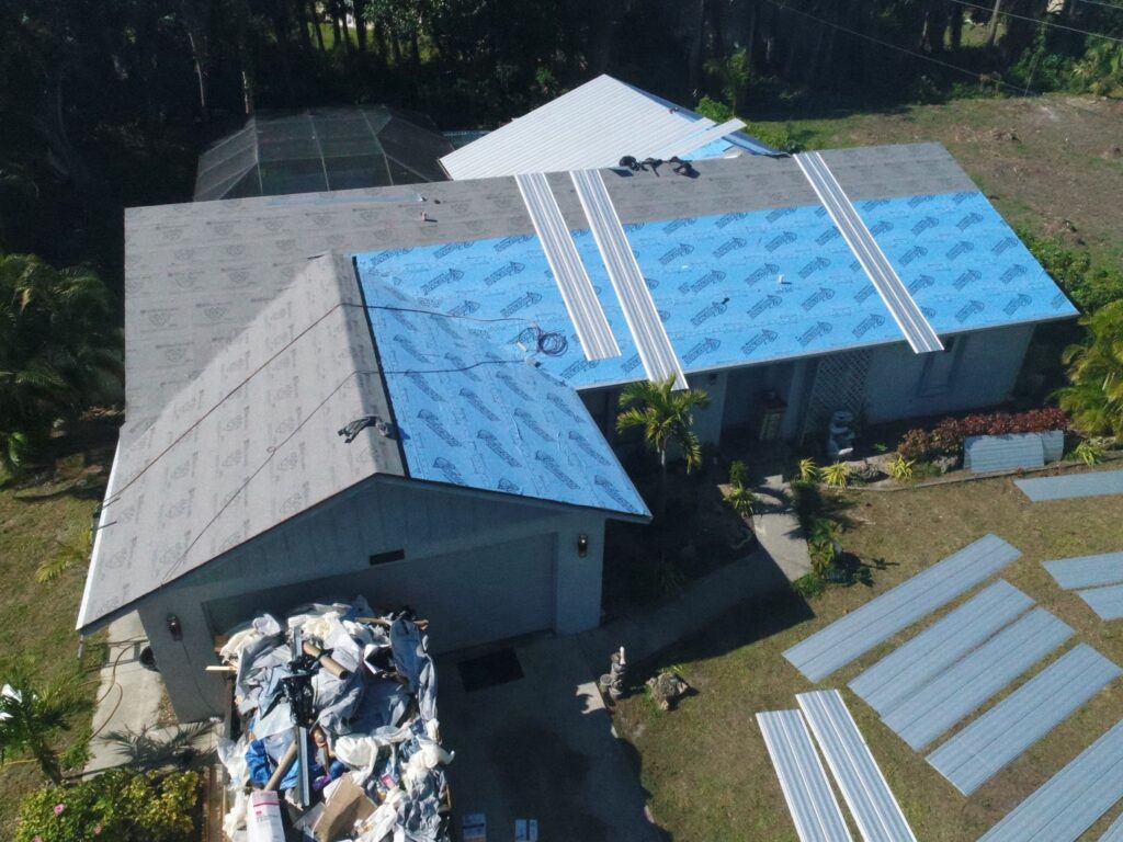 Aerial view of a house undergoing roof repair with a partial roof replacement, showcasing blue underlayment and stacked metal sheets nearby, expertly managed by skilled roofing contractors.