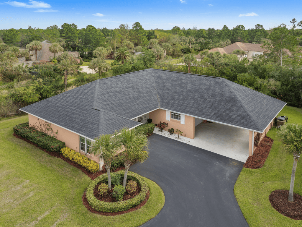 Aerial view of a single-story house with a sleek black metal roofing, driveway, and beautifully landscaped yard featuring trees and shrubs.