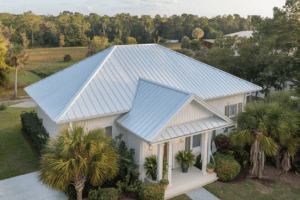 A white house with sleek metal roofing nestles among palm trees and lush greenery, basking under a clear sky.