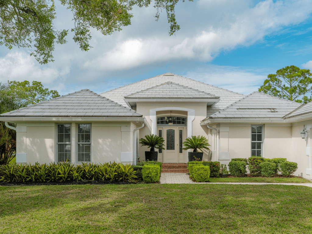 A white single-story house with a tiled roof showcases both elegance and practicality, featuring a landscaped front yard and large entrance under a blue sky. The chosen roof material durability promises longevity, adding lasting value to this charming abode.