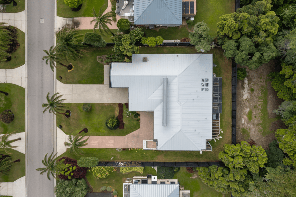 Aerial view of a modern house with a hurricane-proof metal roof, surrounded by trees and a driveway in a suburban neighborhood.