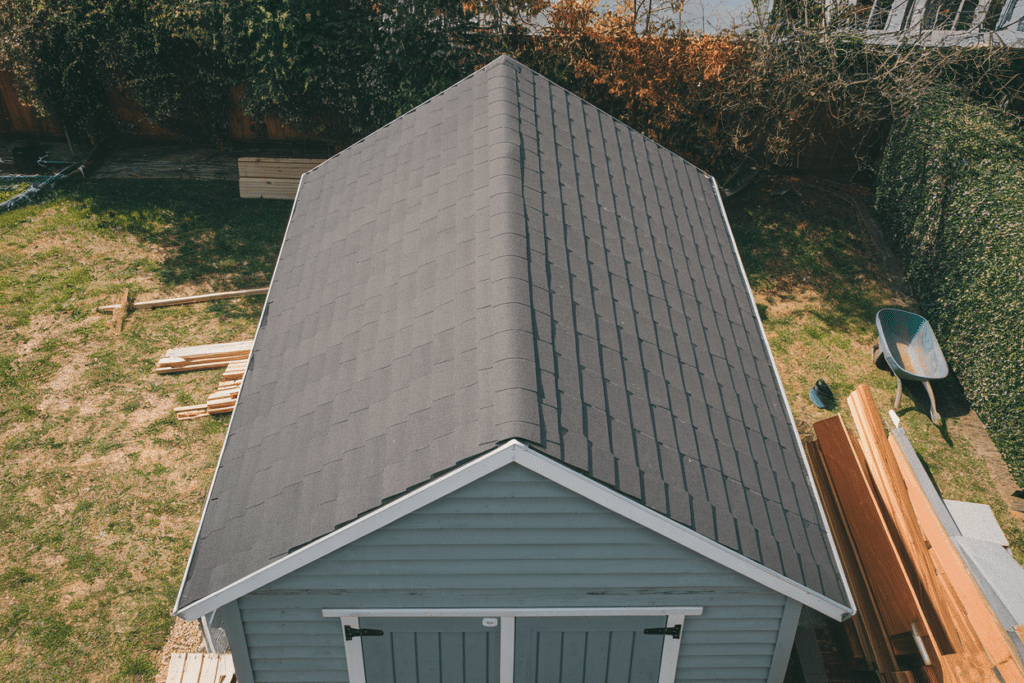 An aerial view reveals a small gray shed with a dark, wind-resistant roof, surrounded by lush grass and scattered construction materials.